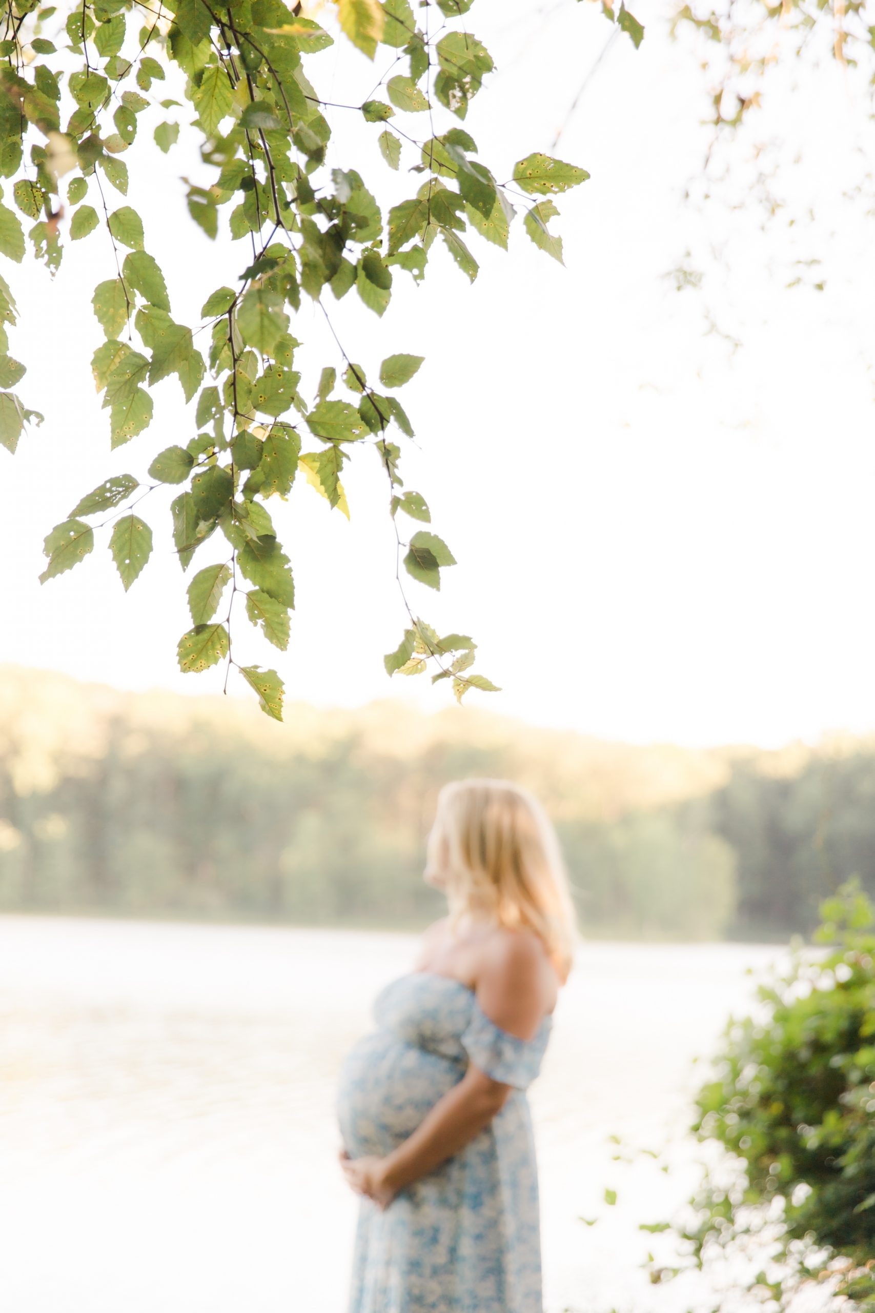 mom-to-be standing by the lake, blurred background with leaves in focus by Northern Virginia Maternity Photographer