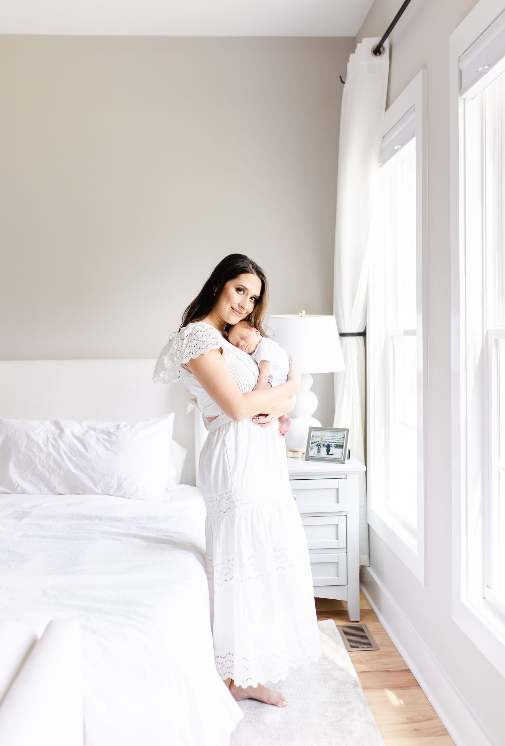 Mom in a long white dress snuggling newborn baby looking at the camera by Northern Virginia Newborn Photographer