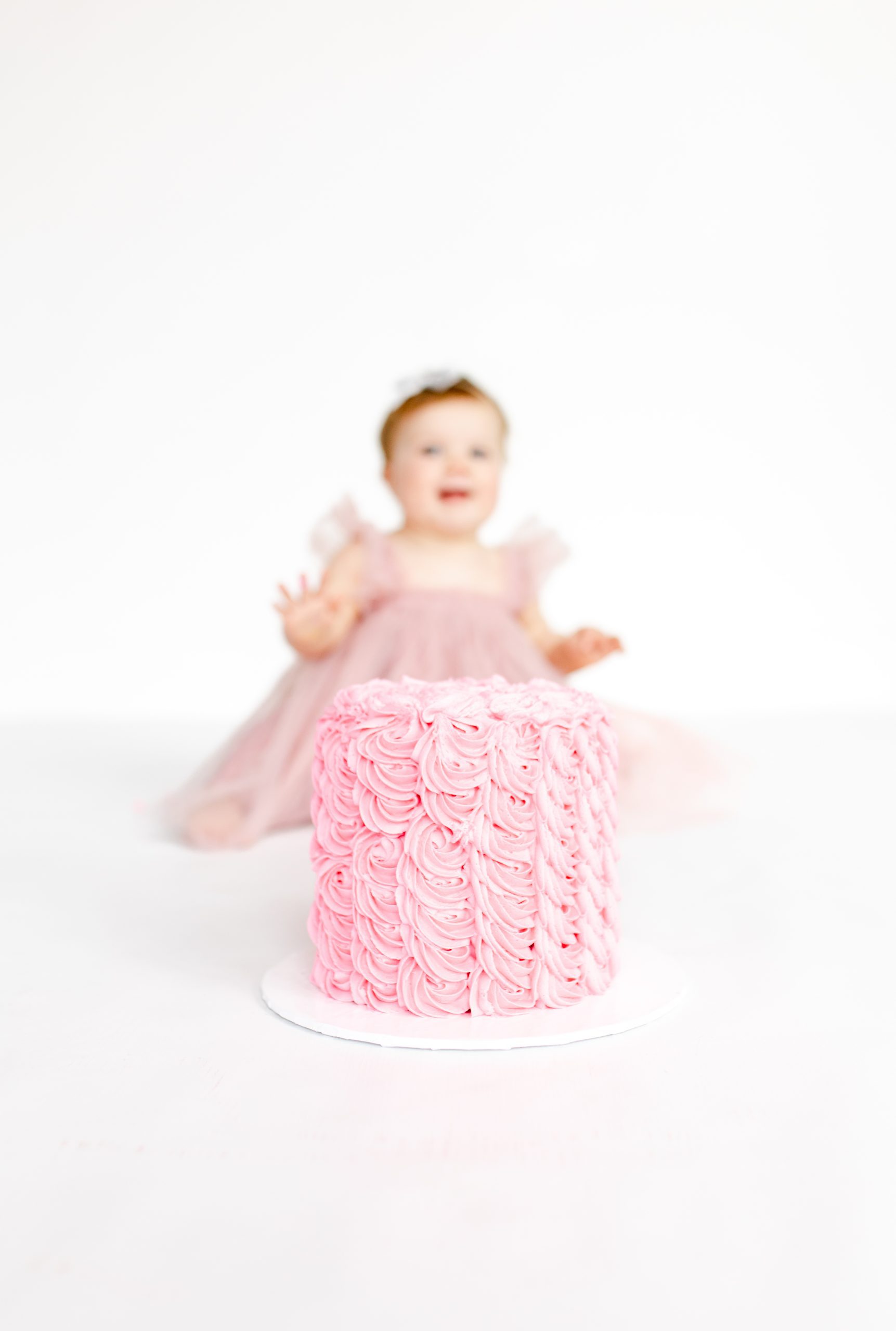 pink smash cake with a baby girl blurred in the background, in a pink dress looking at her smash cake by Northern Virginia Baby Photographer