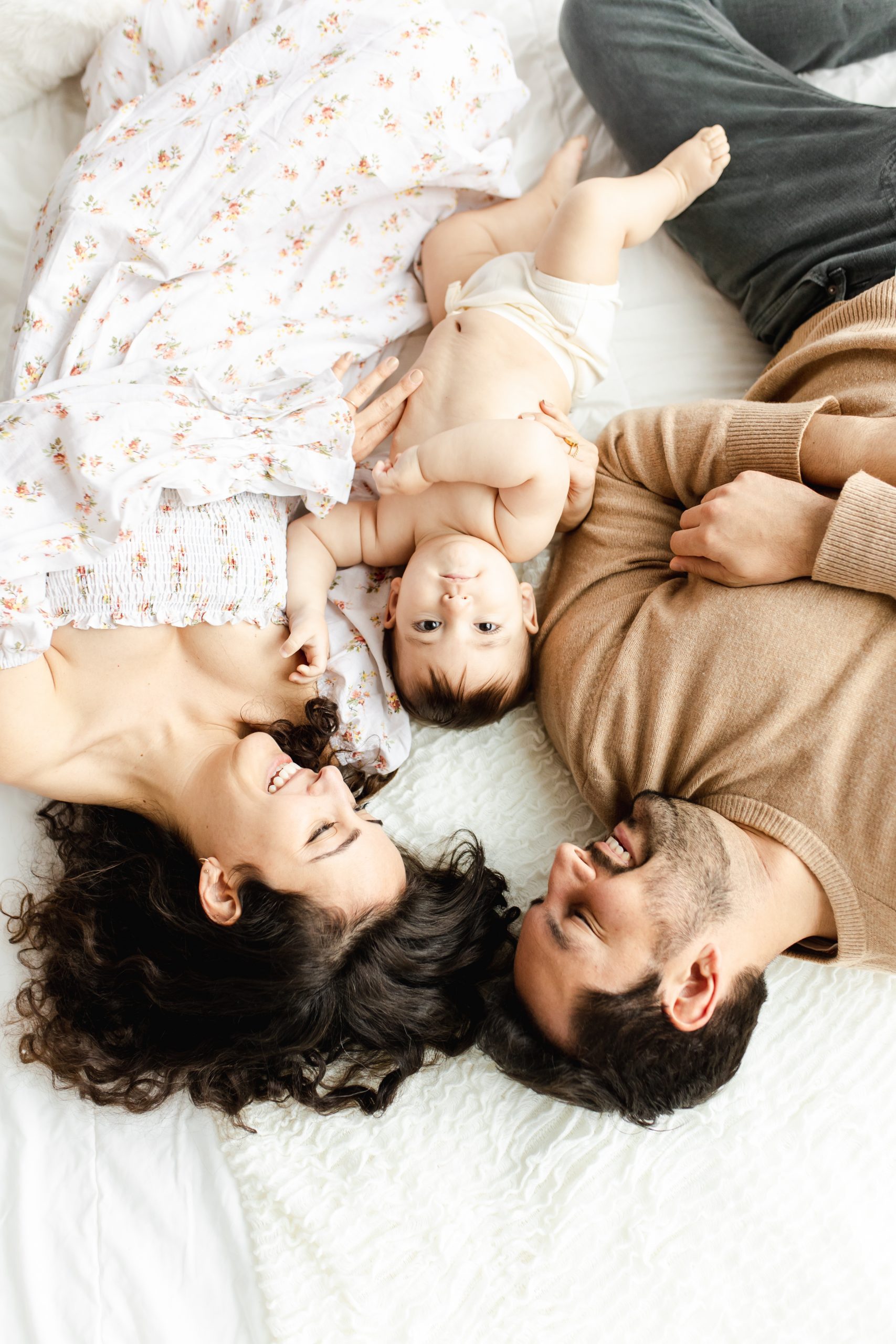 mom, dad and baby laying on a bed, baby looking up at the camera by Northern Virginia Baby Photographer