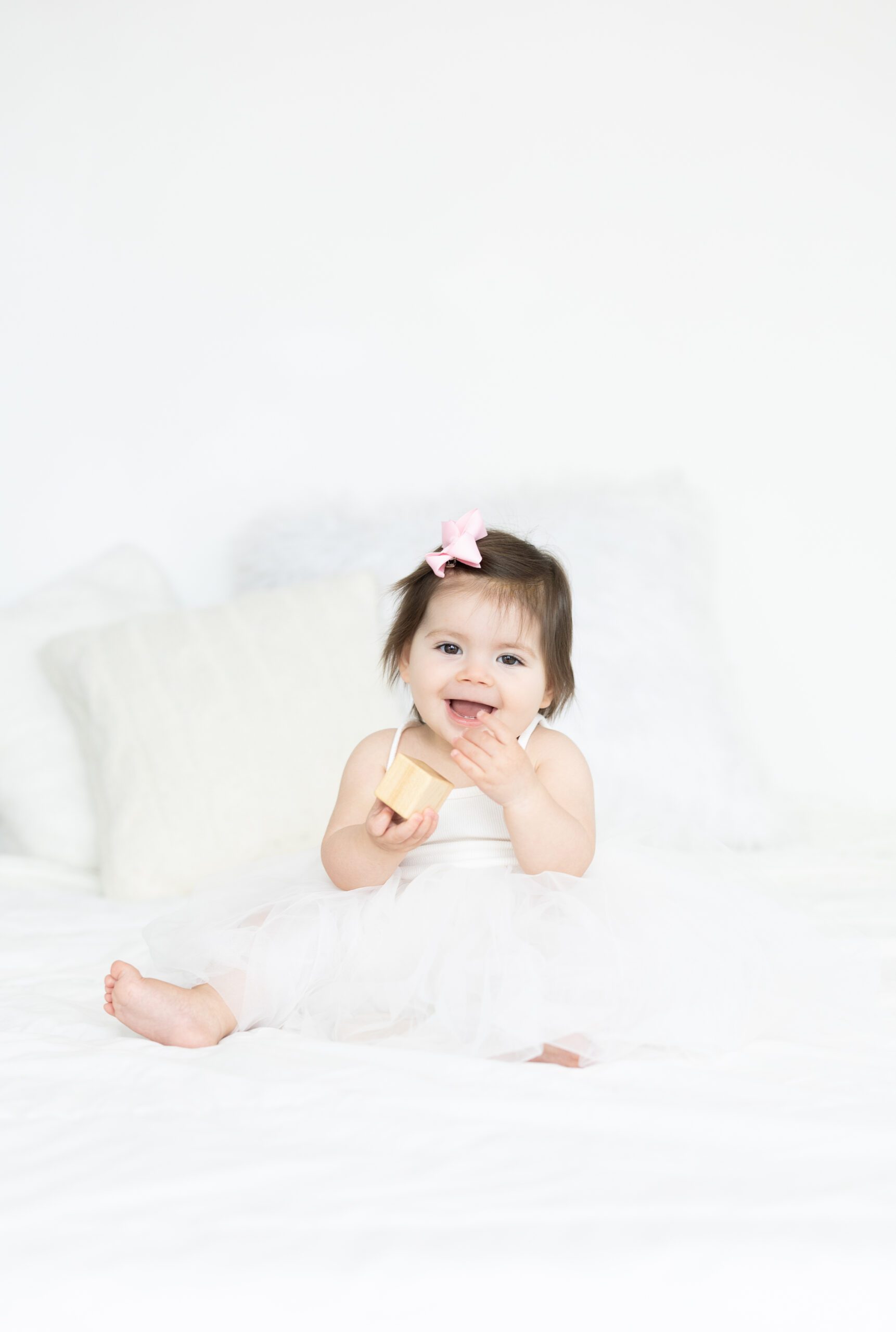 One year old girl in a white dress with a pink bow in an all white studio playing with blocks