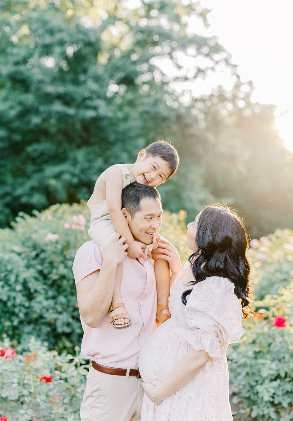 A happy pregnant family plays in a garden with toddler on dad's shoulders