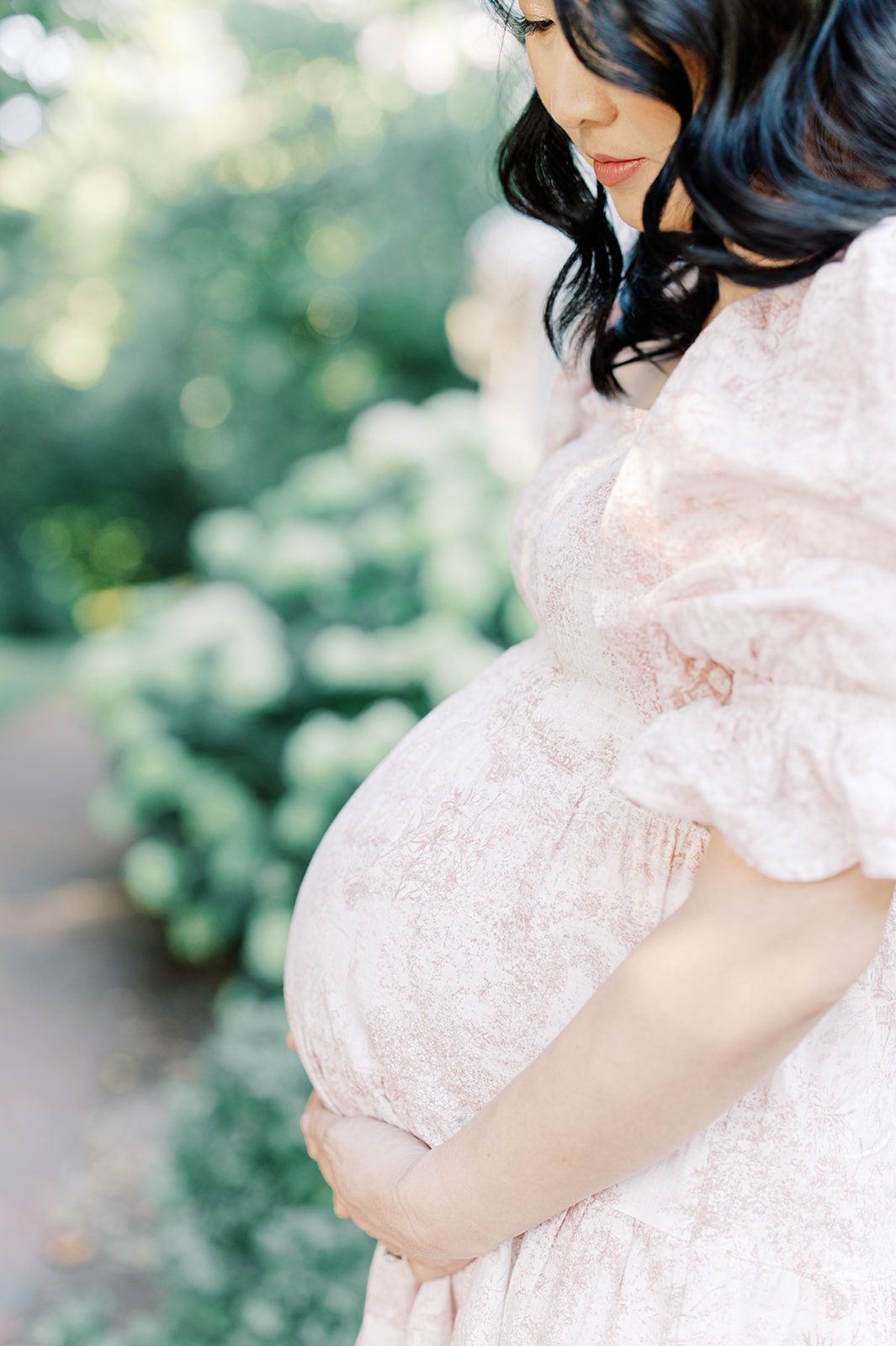A mom to be holds her bump in a pink dress in a garden before visiting baby shower venues in DC