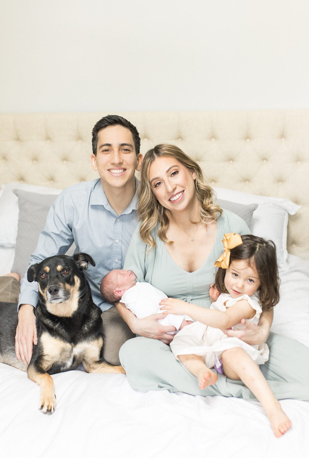 Happy mom and dad sit on a bed holding their newborn, toddler daughter and dog in their laps