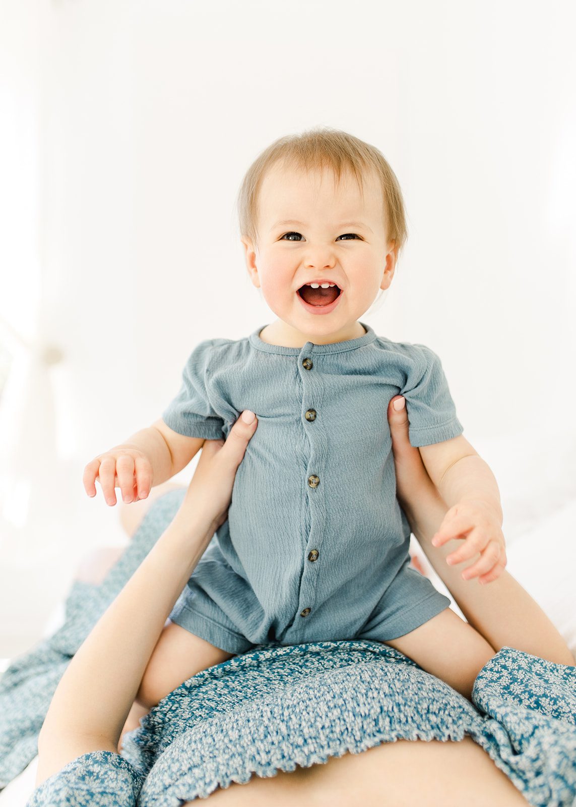 A happy baby plays on mom's belly as she lays on a bed in a studio in a blue dress and blue onesie