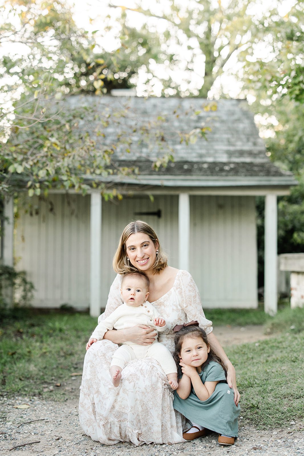 A happy mom kneels with her baby in her lap and toddler daughter at her side for their baby photographer in DC